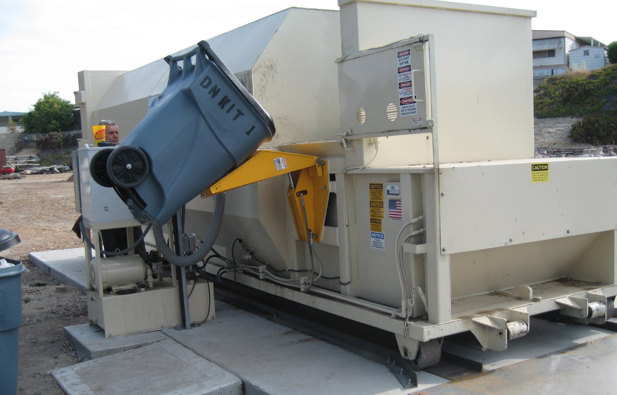 a white horizontal baler pictured outside lifting a trash can preparing to deposit it's contents inside the baler